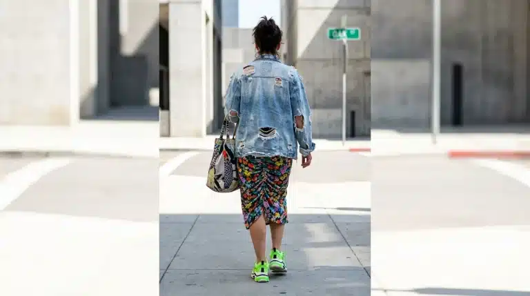 A person walking, viewed from behind, wearing a chaotic outfit of distressed denim, floral print, neon sneakers, and a patterned bag, illustrating common trend-stacking mistakes in outfit inspirations.