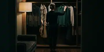 A professional fashion stylist, seen from behind, selecting a versatile dark green silk blouse from a meticulously curated wardrobe, lit by a dramatic single light source, symbolizing strategic seasonal adaptation.
